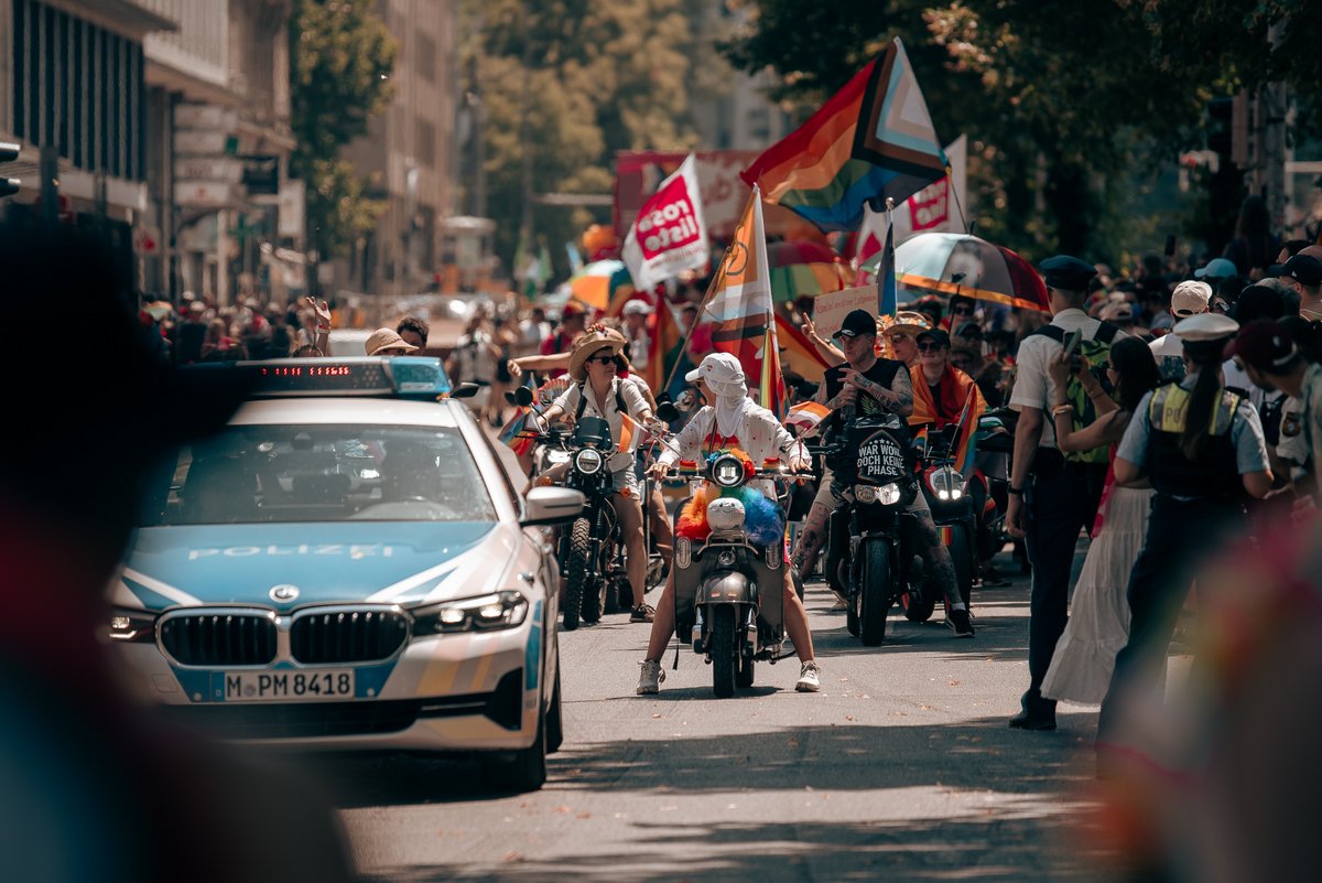 Polizeifahrzeug begleitet die CSD-Parade in München, dahinter fahren Motorräder mit Regenbogen- und Progress-Pride-Flaggen.