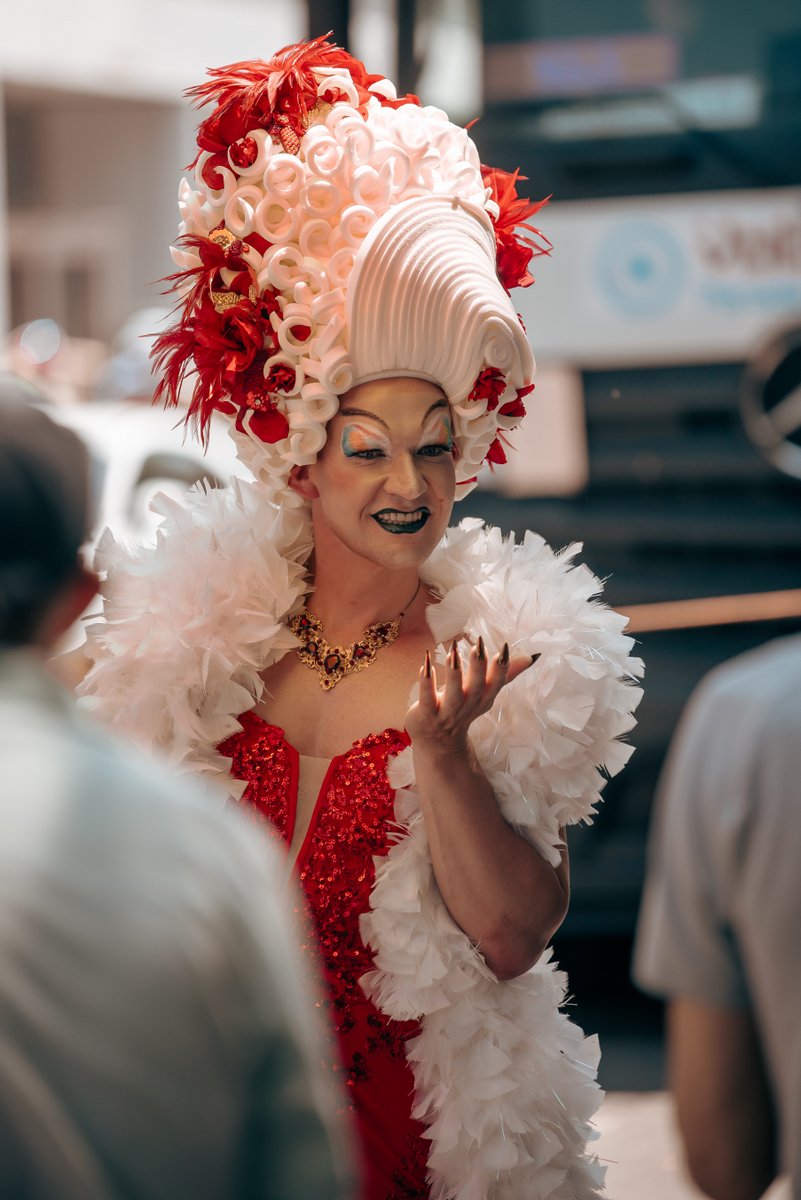 Drag Artist in aufwendigem rot-wei?m Kost? und gro?r Per?ke begr?t die Zuschauerinnen und Zuschauer am Stra?nrand.