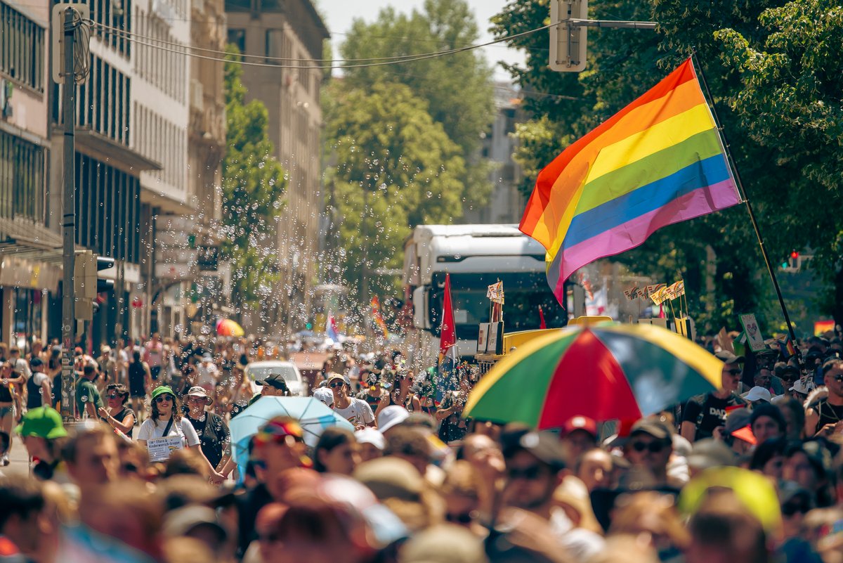Gro? Regenbogenflagge weht ?er der dicht gef?lten CSD-Parade in München an einem hei?n Sommertag.