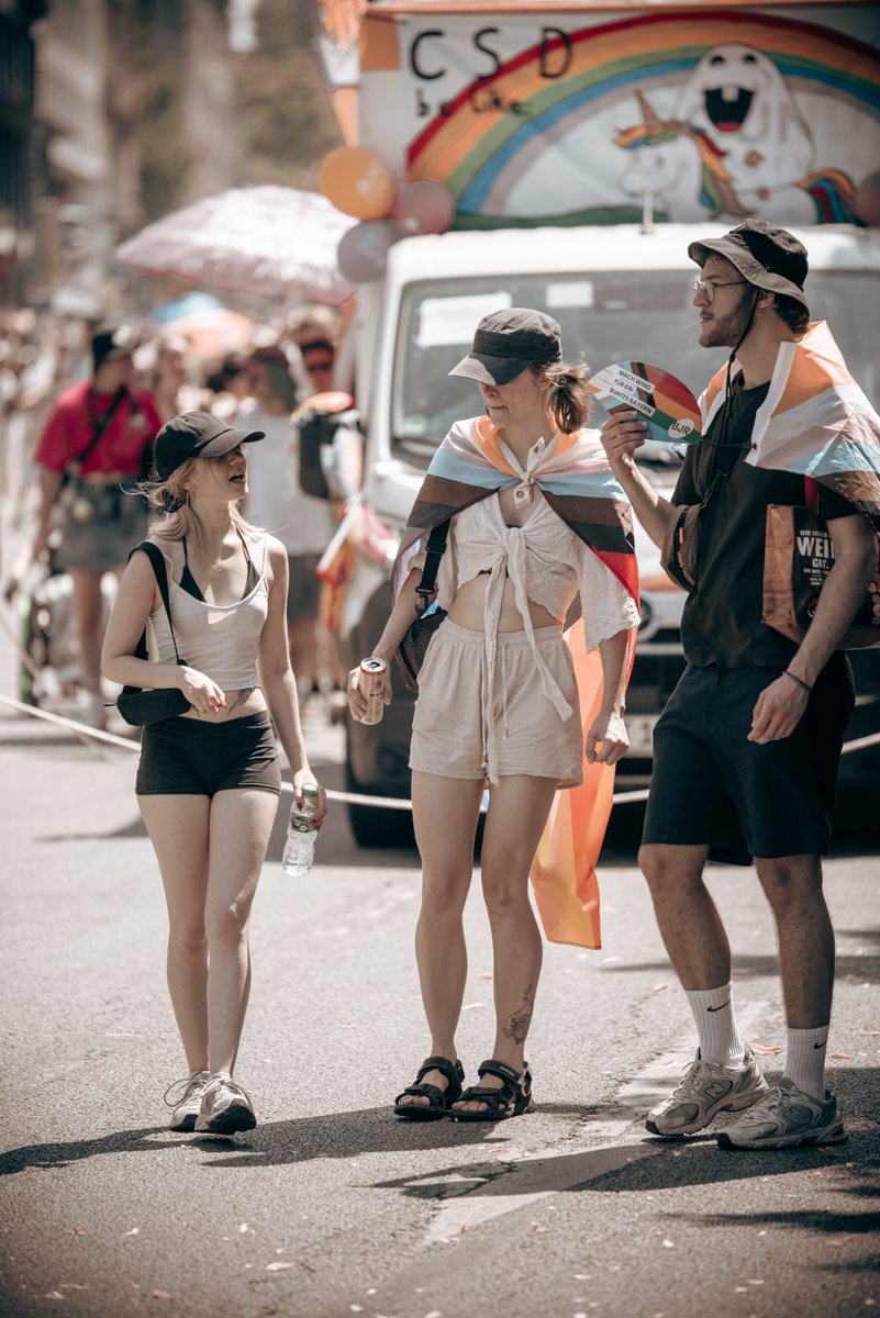 Drei junge Teilnehmende mit Pride-Flaggen und F?hern stehen vor einem bunt geschm?kten CSD-Fahrzeug.