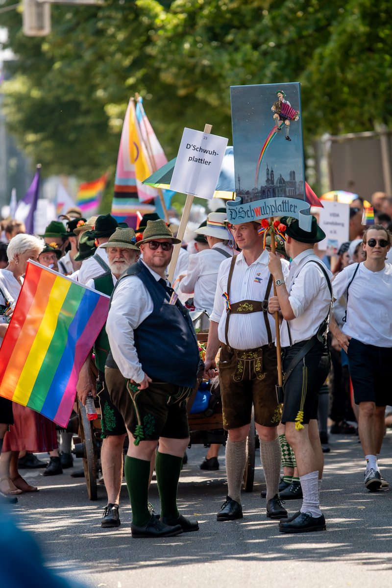 Gruppe in Tracht und sommerlicher Kleidung steht mit Regenbogenflaggen am Rand der CSD-Parade.