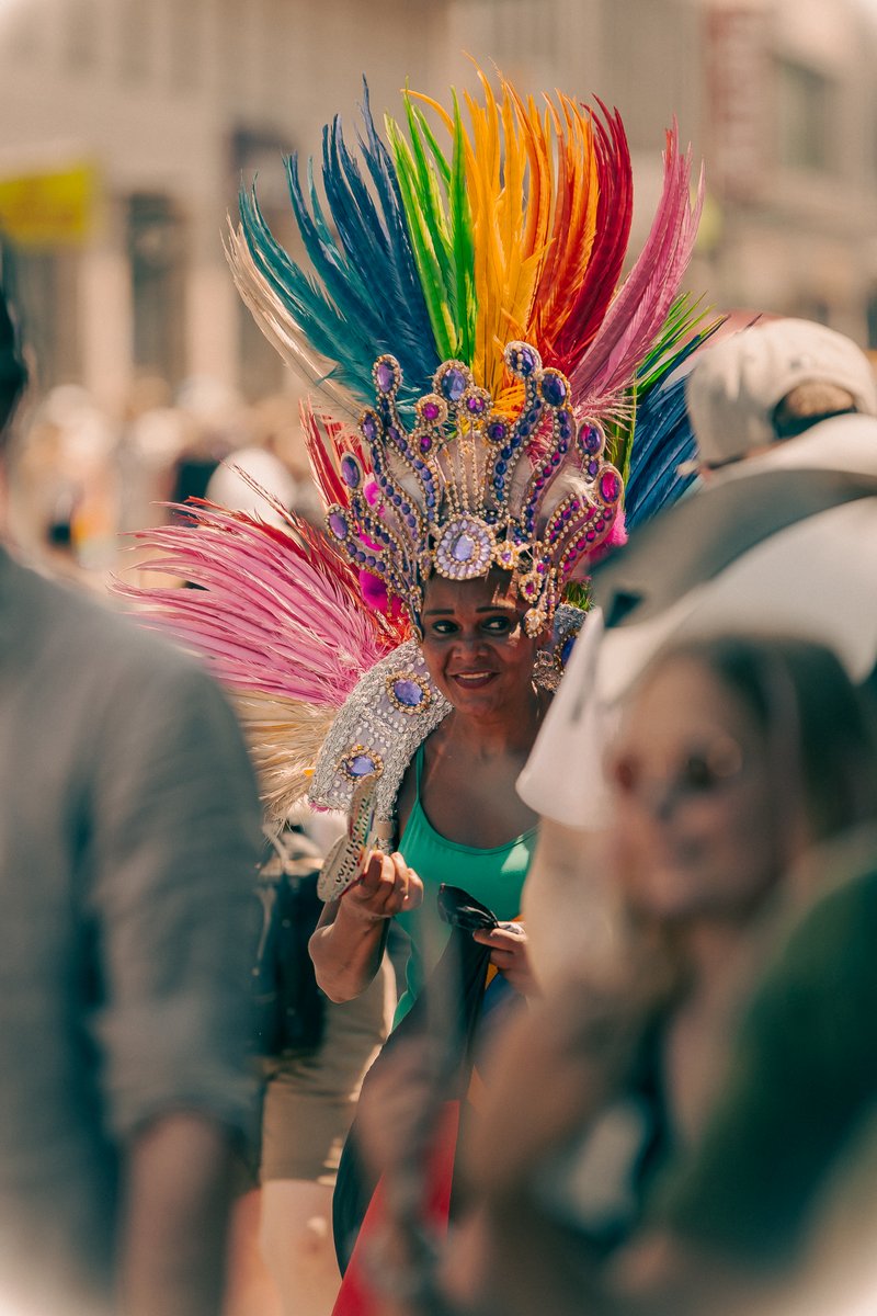 Person mit buntem Federkopfschmuck steht dicht zwischen Zuschauenden und Pride-Teilnehmenden.
