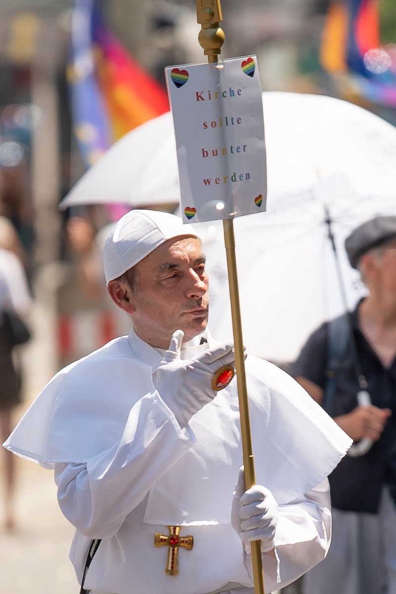 Person im wei?n Papst-Kost? tr?t ein Schild mit Regenbogenbotschaft durch die Parade.