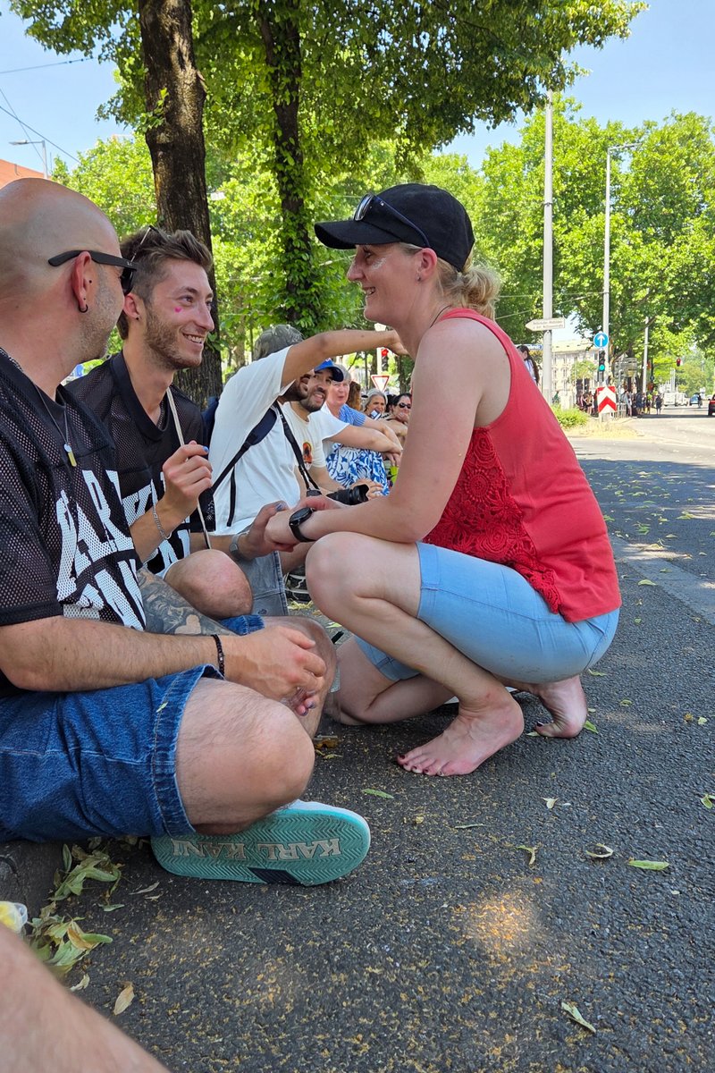 Drei Personen sitzen im Schatten am Stra?nrand und unterhalten sich w?rend einer Pause vom CSD.