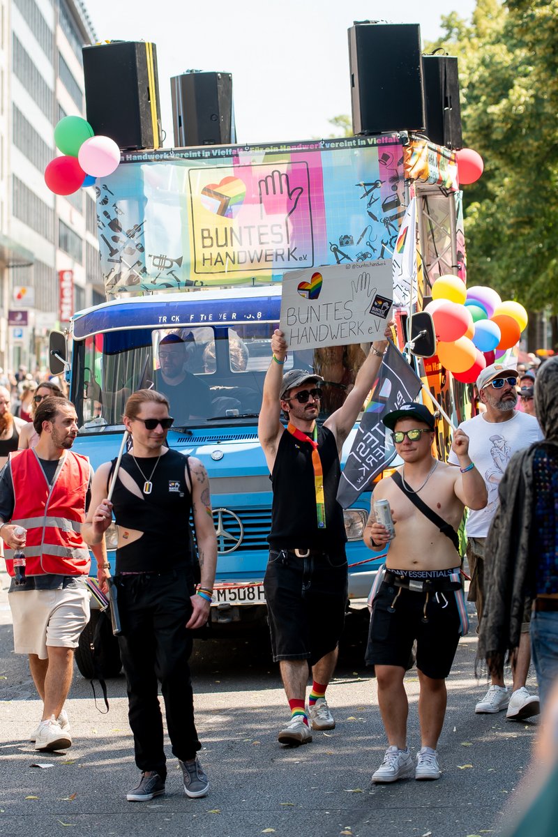 Zwei Personen gehen vor einem Paradewagen mit Ballons und Regenbogenmotiven durch die Parade.