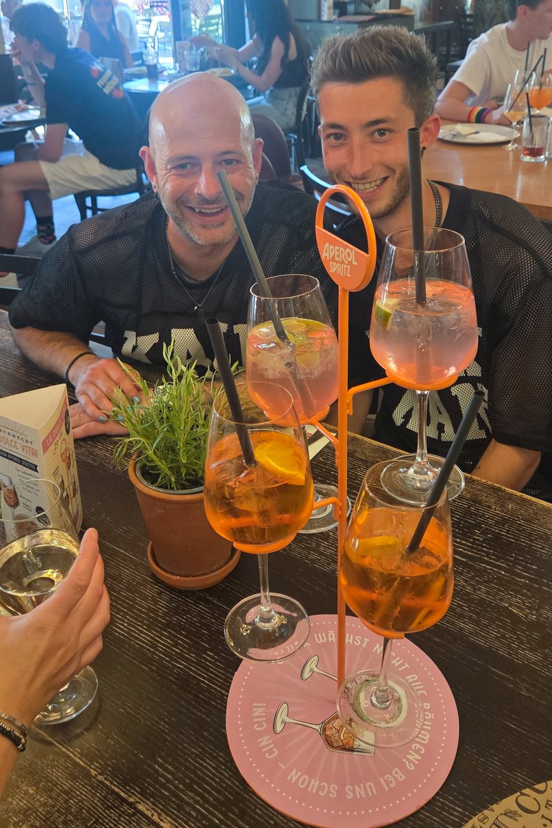 Zwei M?ner sitzen mit gro?n Aperol-Gl?ern im Restaurant und lachen in die Kamera.